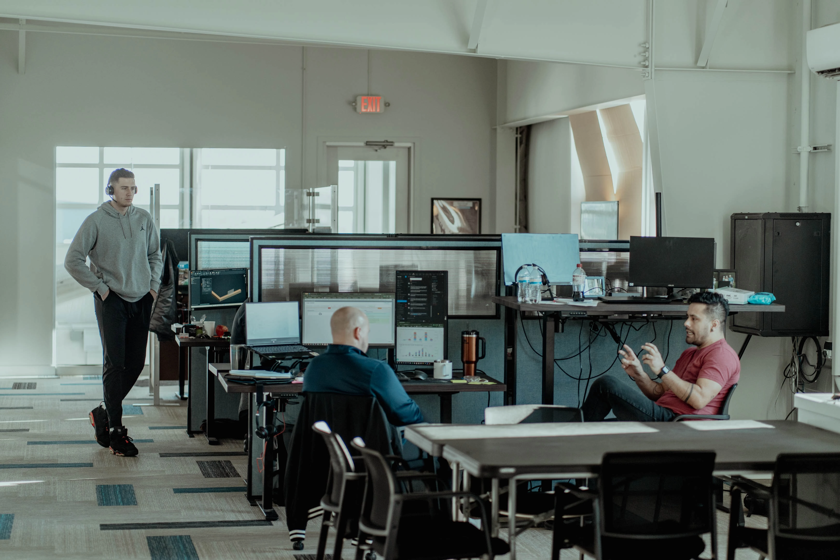 Modern office space with three men working and interacting; one man in a red shirt gestures while seated, another man with a bald head faces multiple monitors, and a third man wearing a gray hoodie and headphones stands nearby.