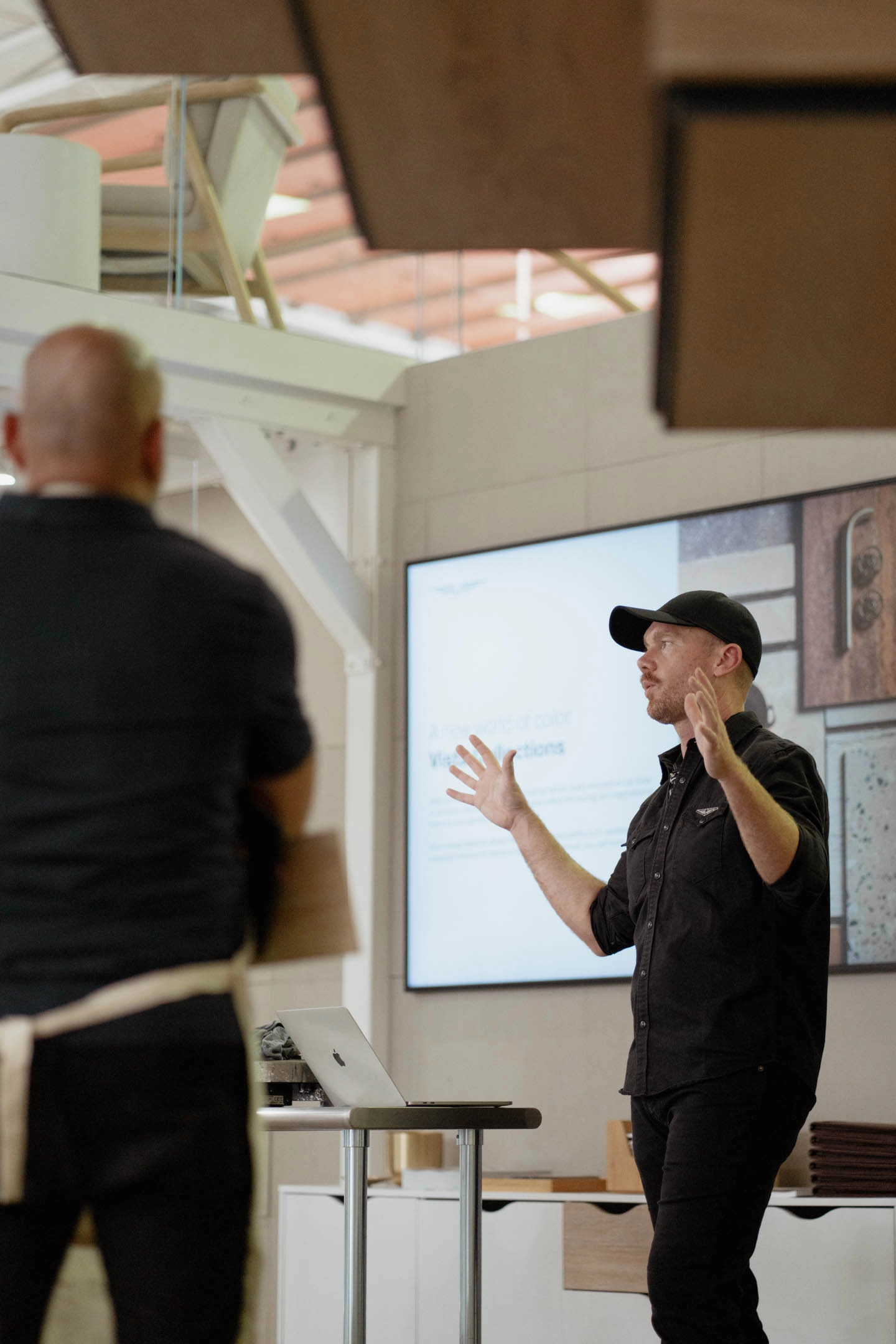At an architect event, a presenter gestures beside a screen below a glass-railed mezzanine