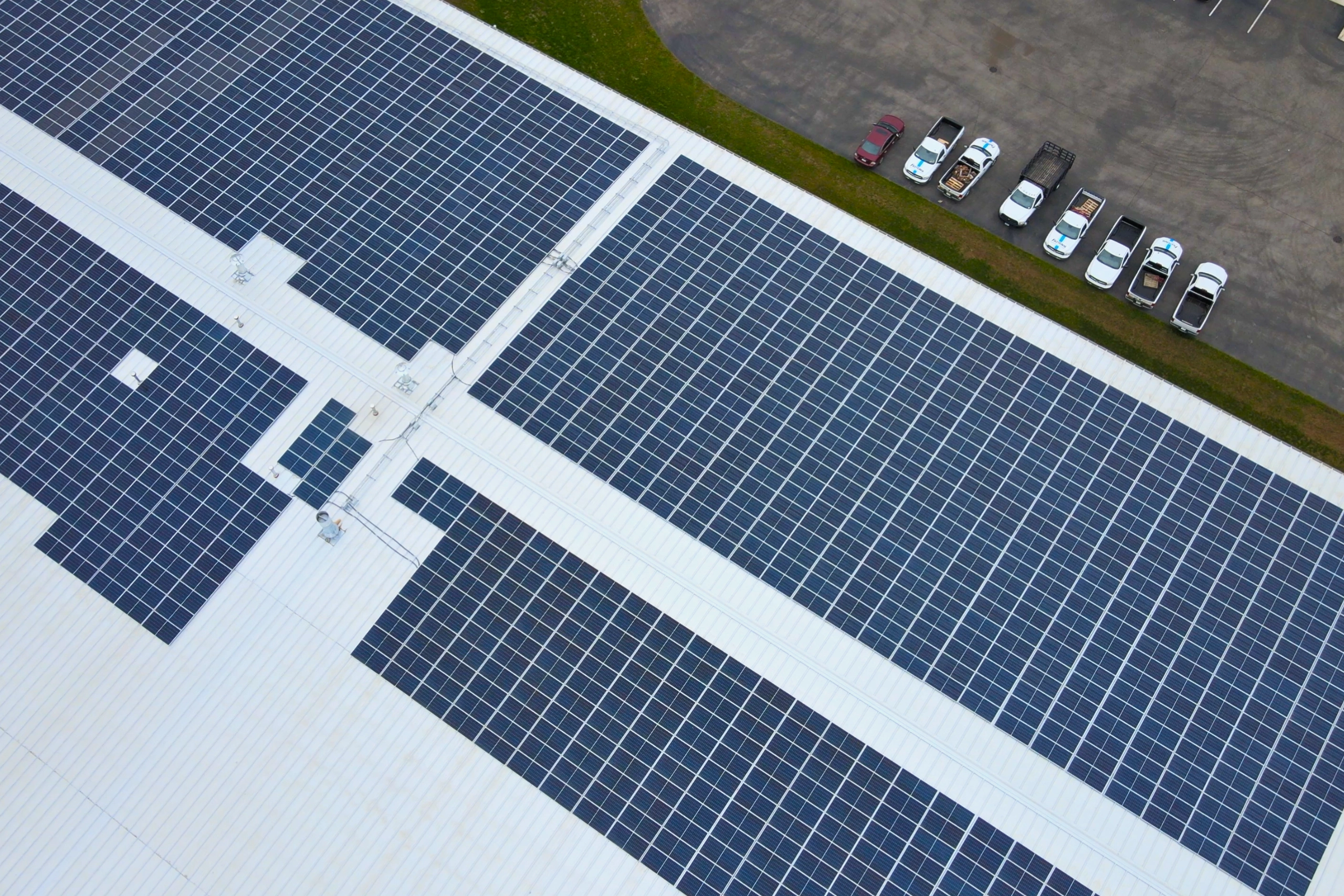 Aerial view of a large white industrial roof covered with multiple blue solar panels arranged in rows, with a row of parked vehicles on the adjacent concrete surface.