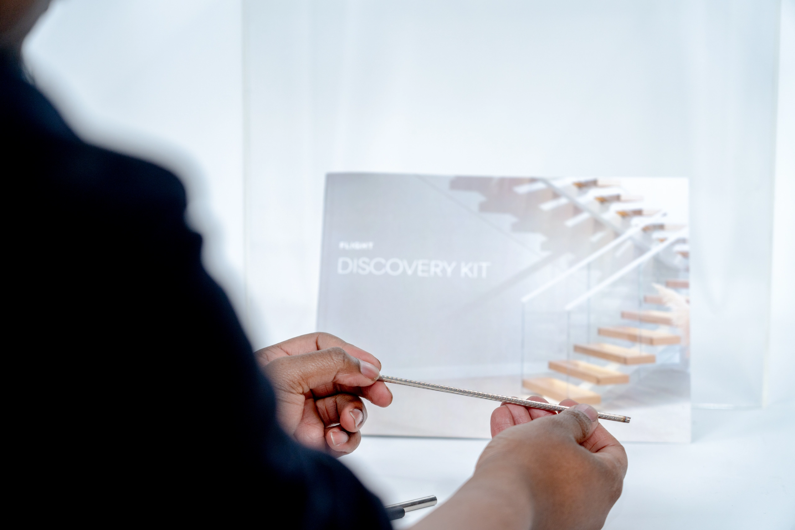 Person holding and examining a thin metallic rod in front of a blurred booklet titled 'Discovery Kit' with an image of a modern staircase.