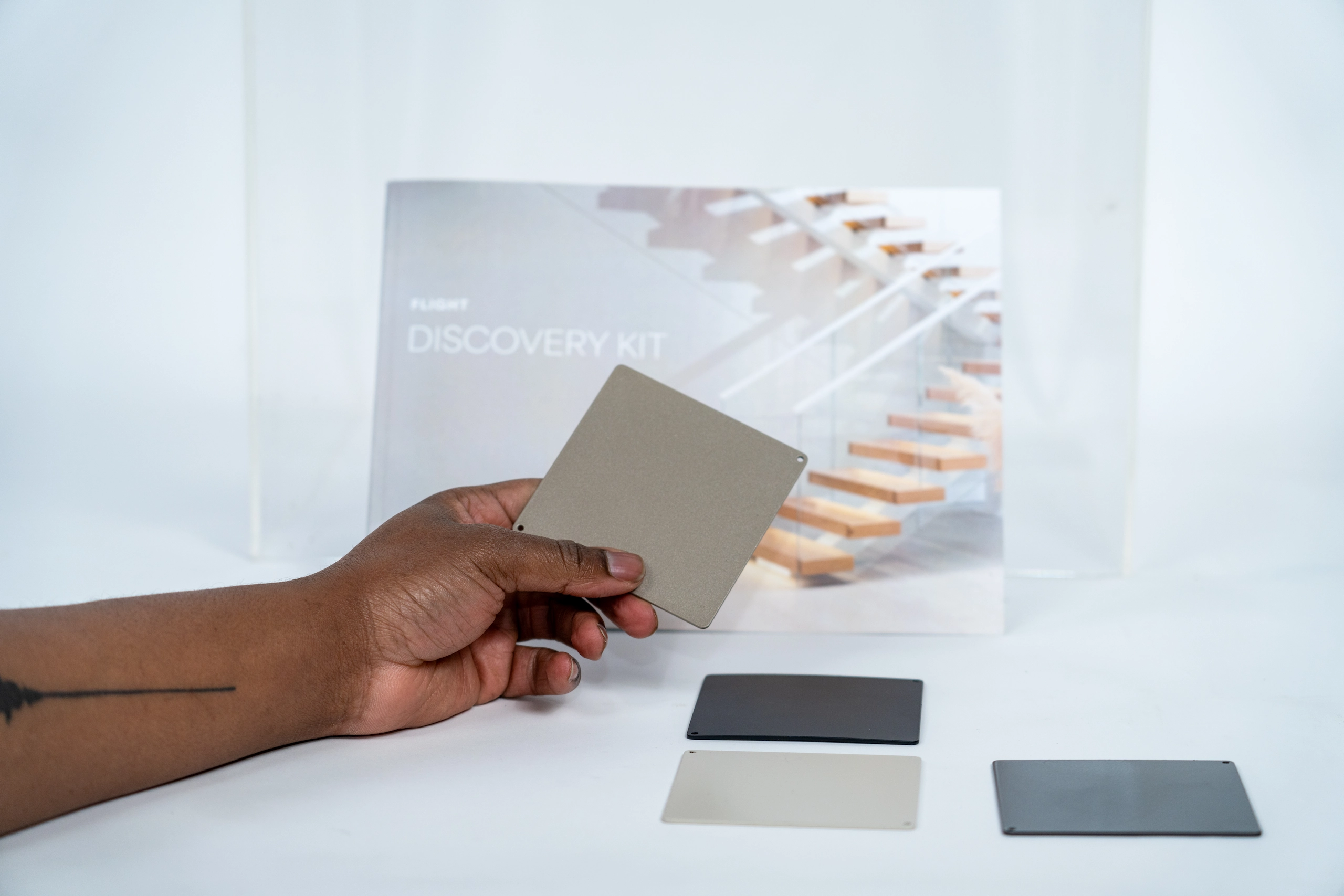 Hand holding a square metallic sample card above three other sample cards in black, gray, and beige on a white surface, with a booklet titled 'Flight Discovery Kit' and an image of a staircase in the background.