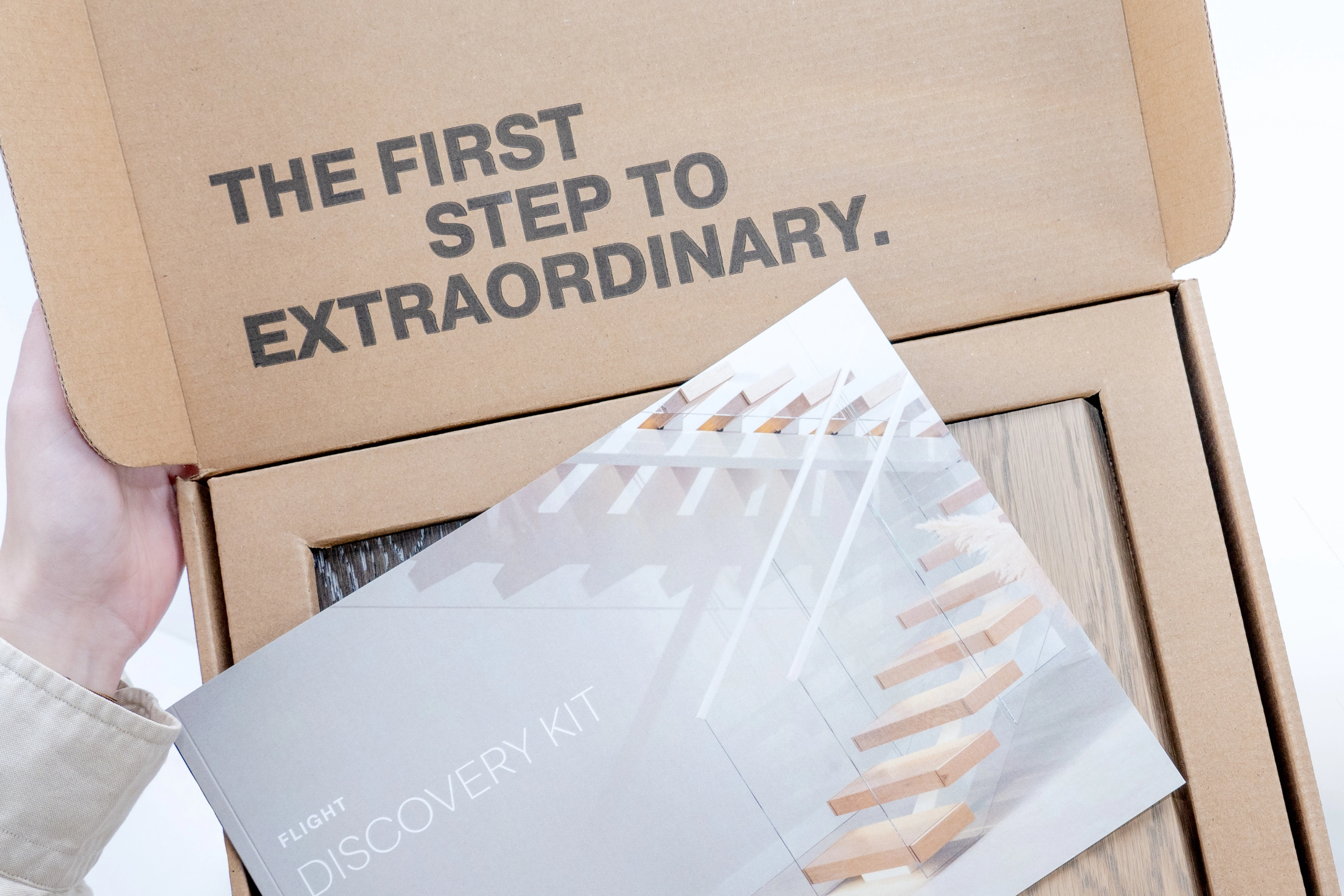 Opened cardboard box held by a hand with text inside the lid reading 'THE FIRST STEP TO EXTRAORDINARY.' A booklet titled 'FLIGHT DISCOVERY KIT' showing a modern glass staircase rests on top of a wood-textured product inside the box.