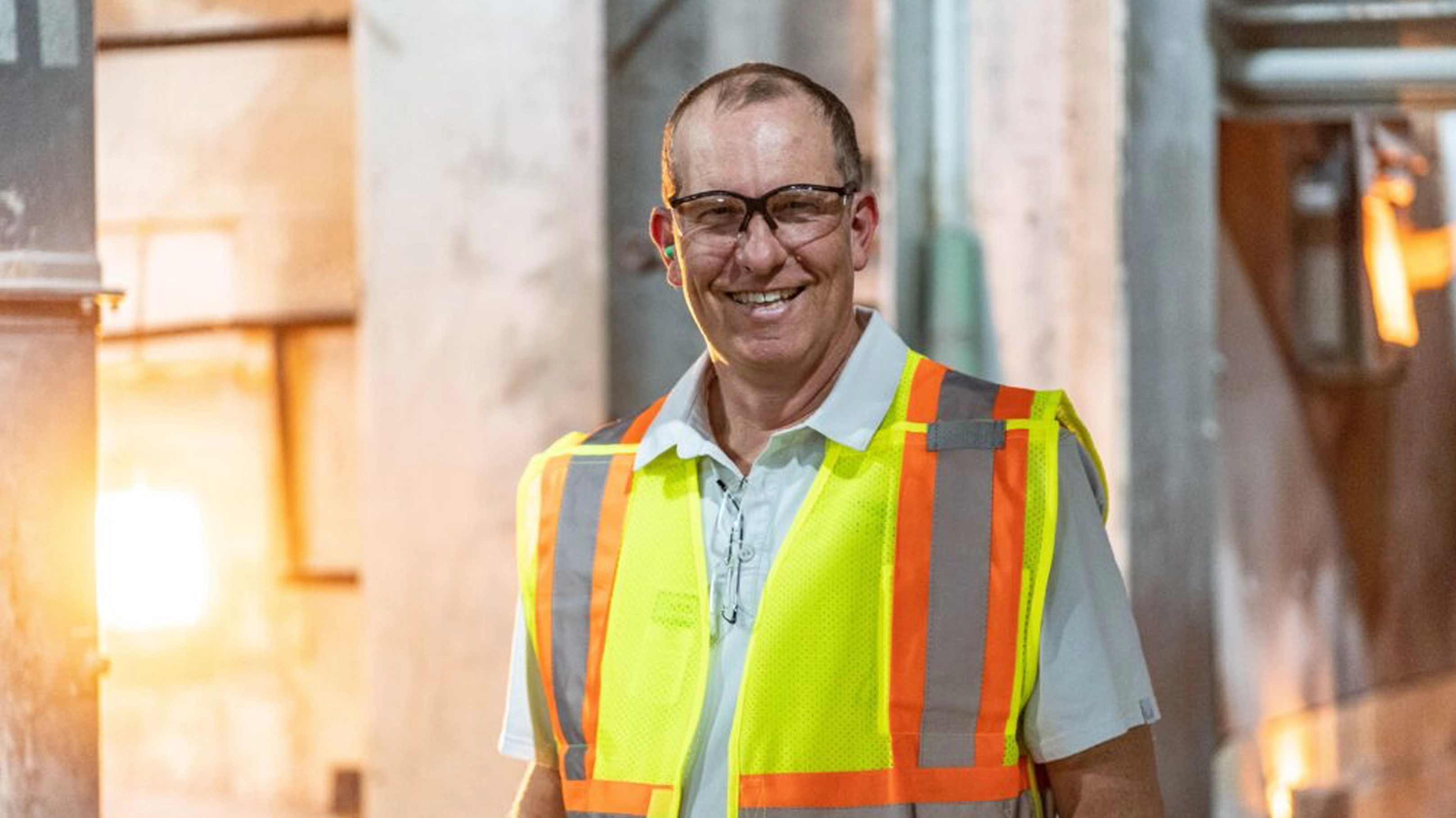 Smiling middle-aged man wearing safety glasses and a high-visibility safety vest in an industrial or construction setting.
