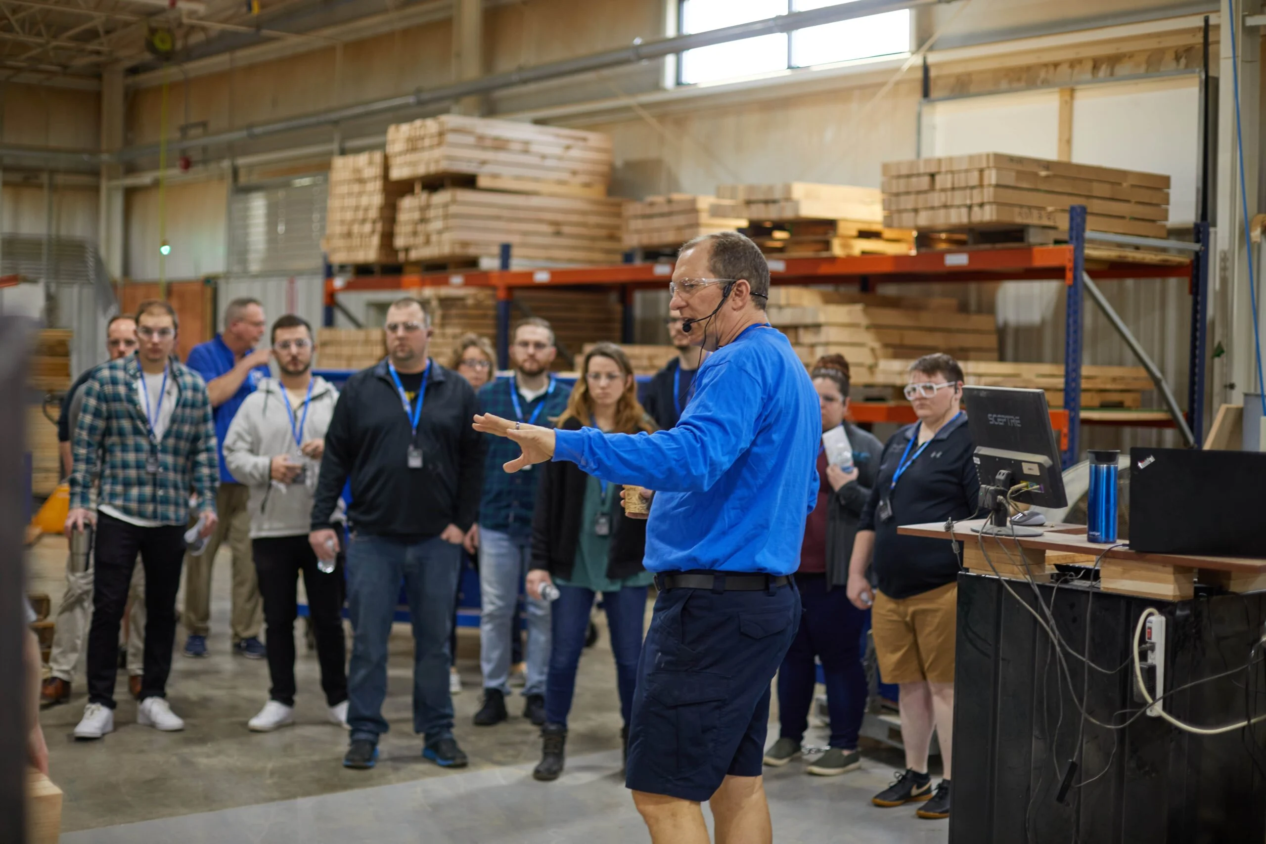 A man wearing a blue long-sleeve shirt, shorts, safety glasses, and a headset speaks to a group of people in a warehouse with stacked wood pallets in the background. The group listens attentively, some holding water bottles, and many wear safety glasses and lanyards with badges.