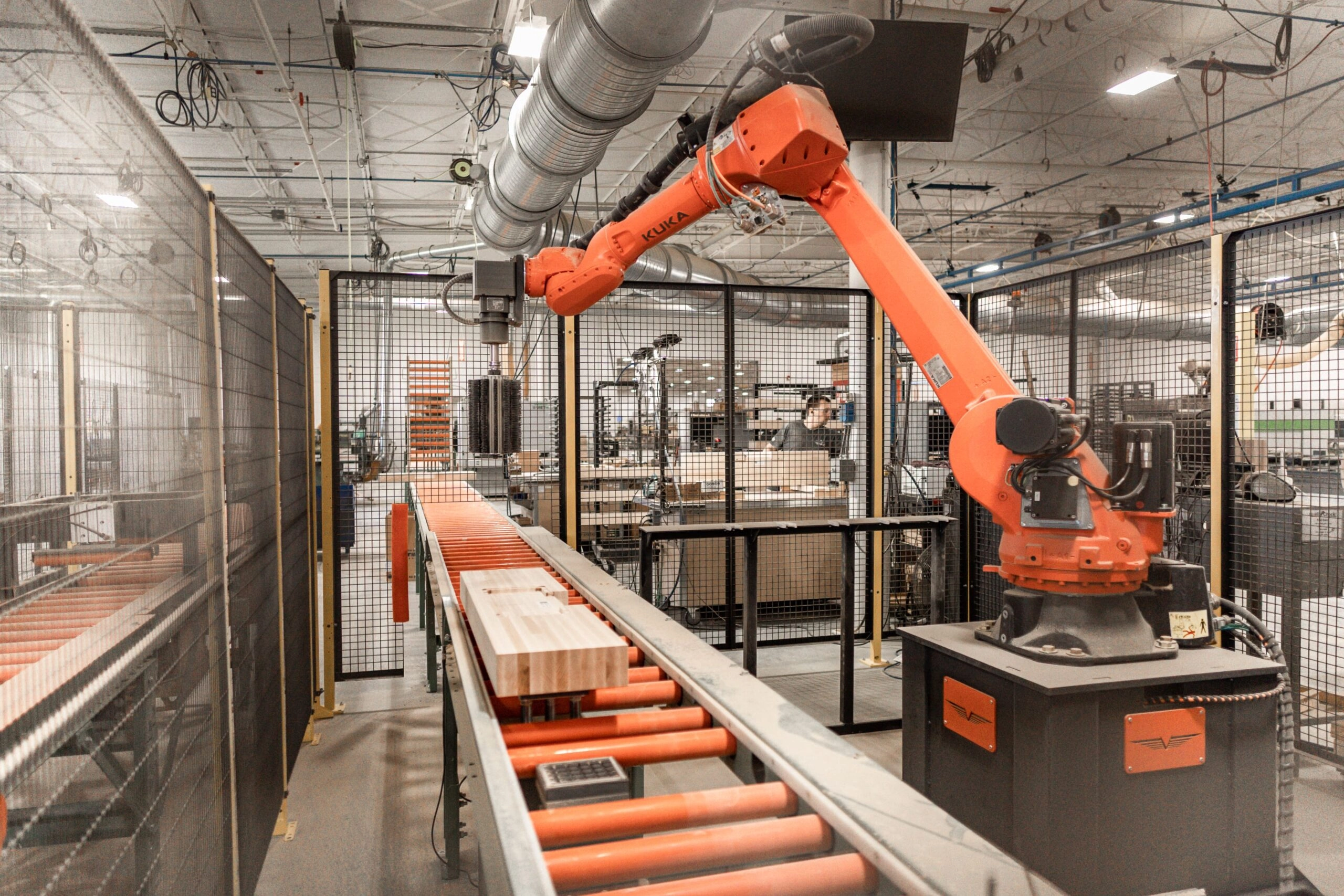 An orange KUKA robotic arm working in a wood production facility, handling wooden blocks on a conveyor belt inside a fenced-off area.