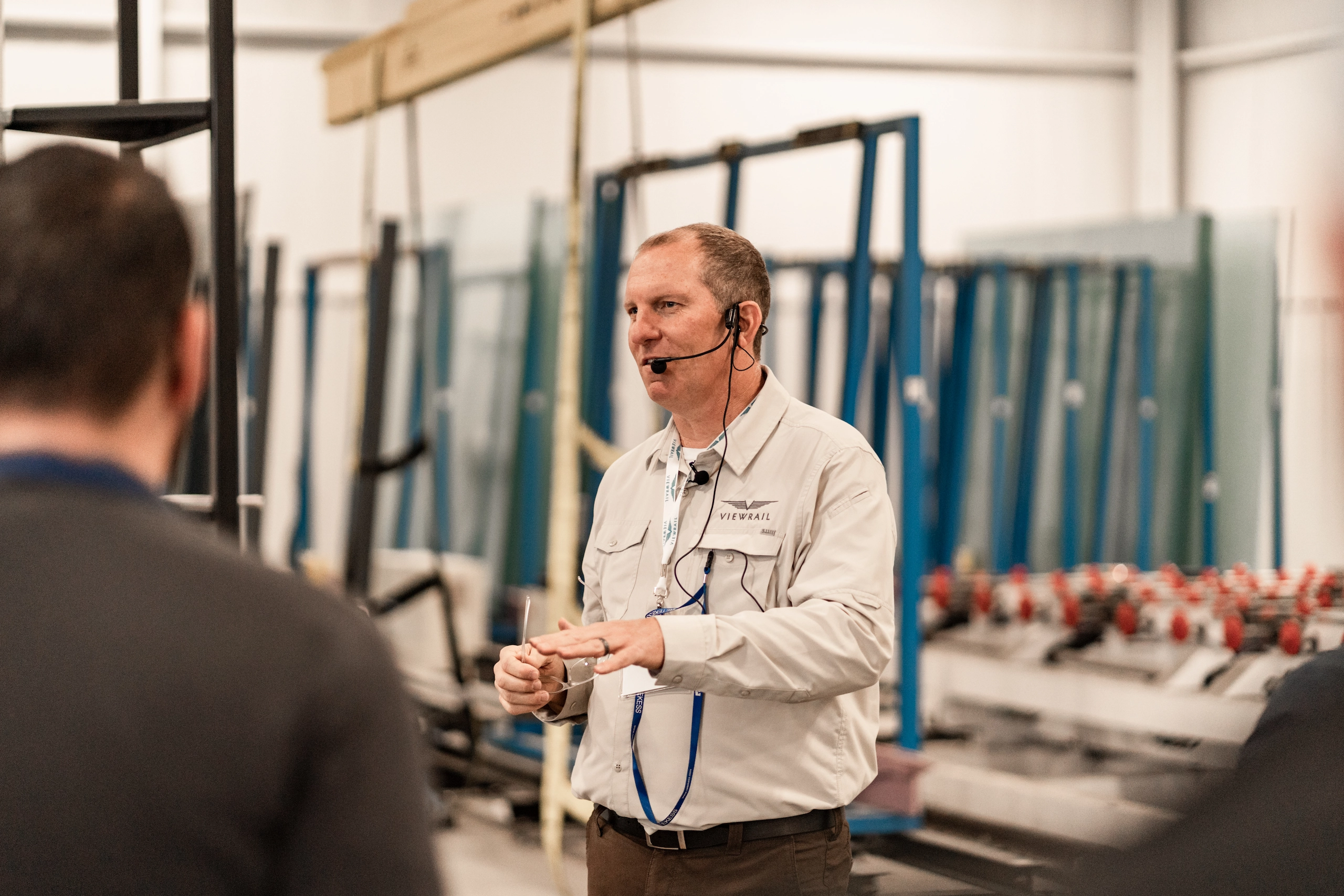 A man wearing a beige Viewrail uniform shirt and headset speaks while holding glasses, standing in an industrial setting with glass panels and equipment racks behind him, facing two blurred individuals in the foreground.