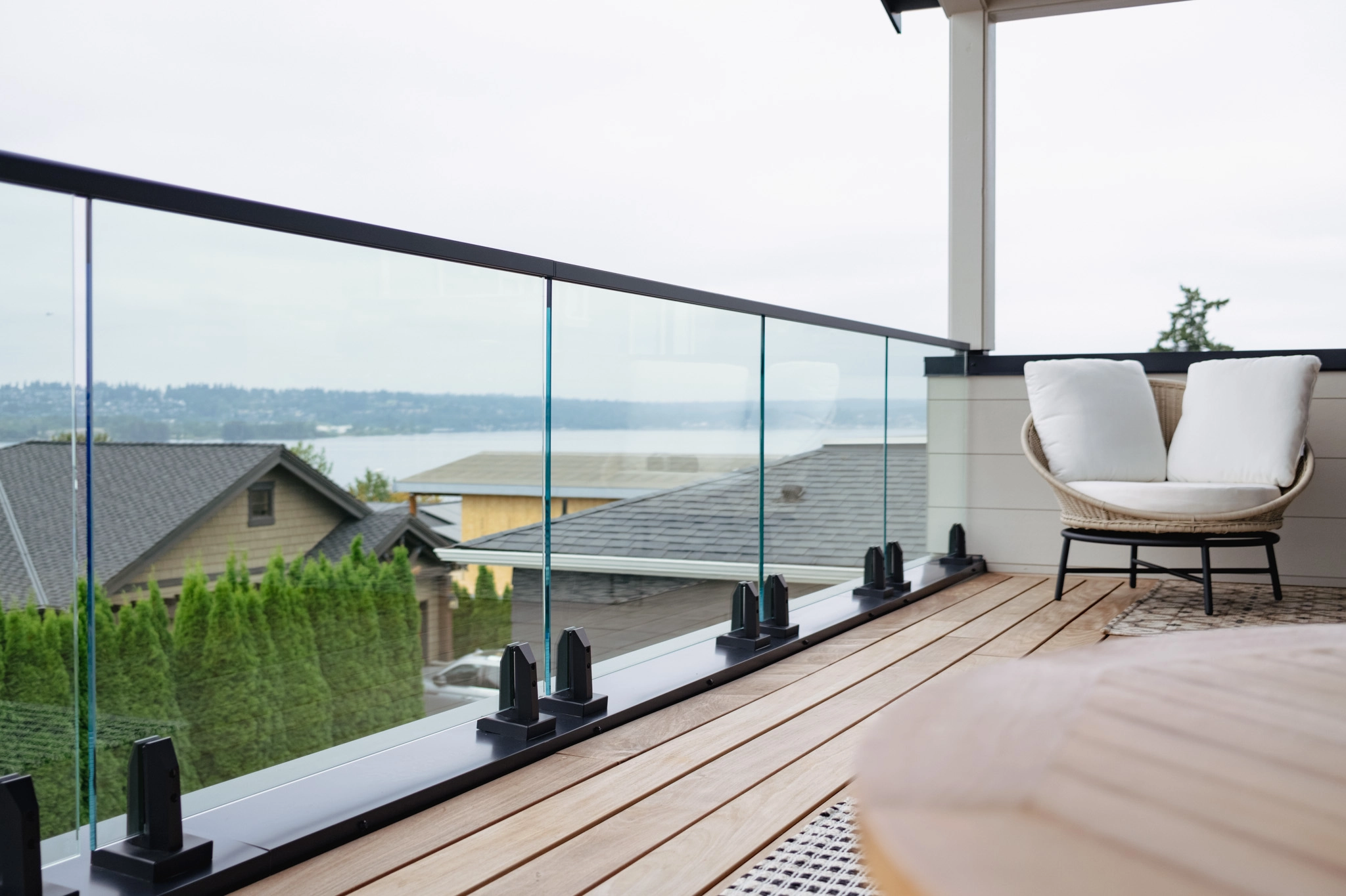 Standard glass panels on black spigots rail a deck balcony above rooftops and a grey bay