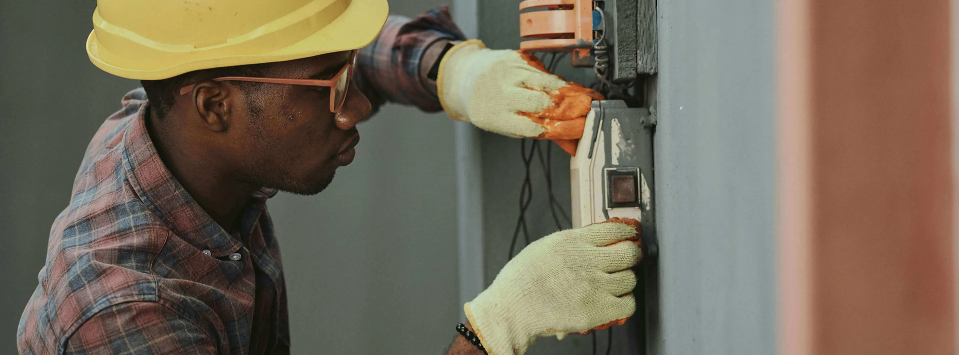 Electrician in yellow hard hat and gloves working on electrical switch on a wall.