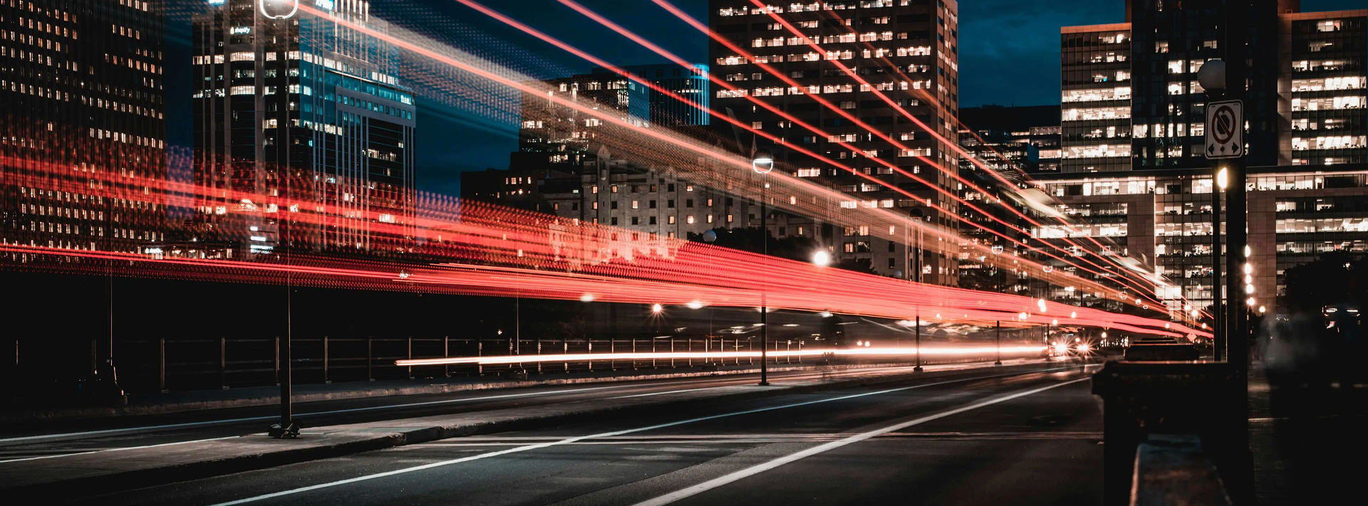 Night cityscape with long-exposure light trails from vehicles on a street among illuminated buildings.