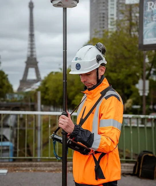 Un homme portant un casque blanc et une veste de sécurité orange réfléchissante utilise un équipement de mesure en plein air avec la Tour Eiffel floue en arrière-plan.