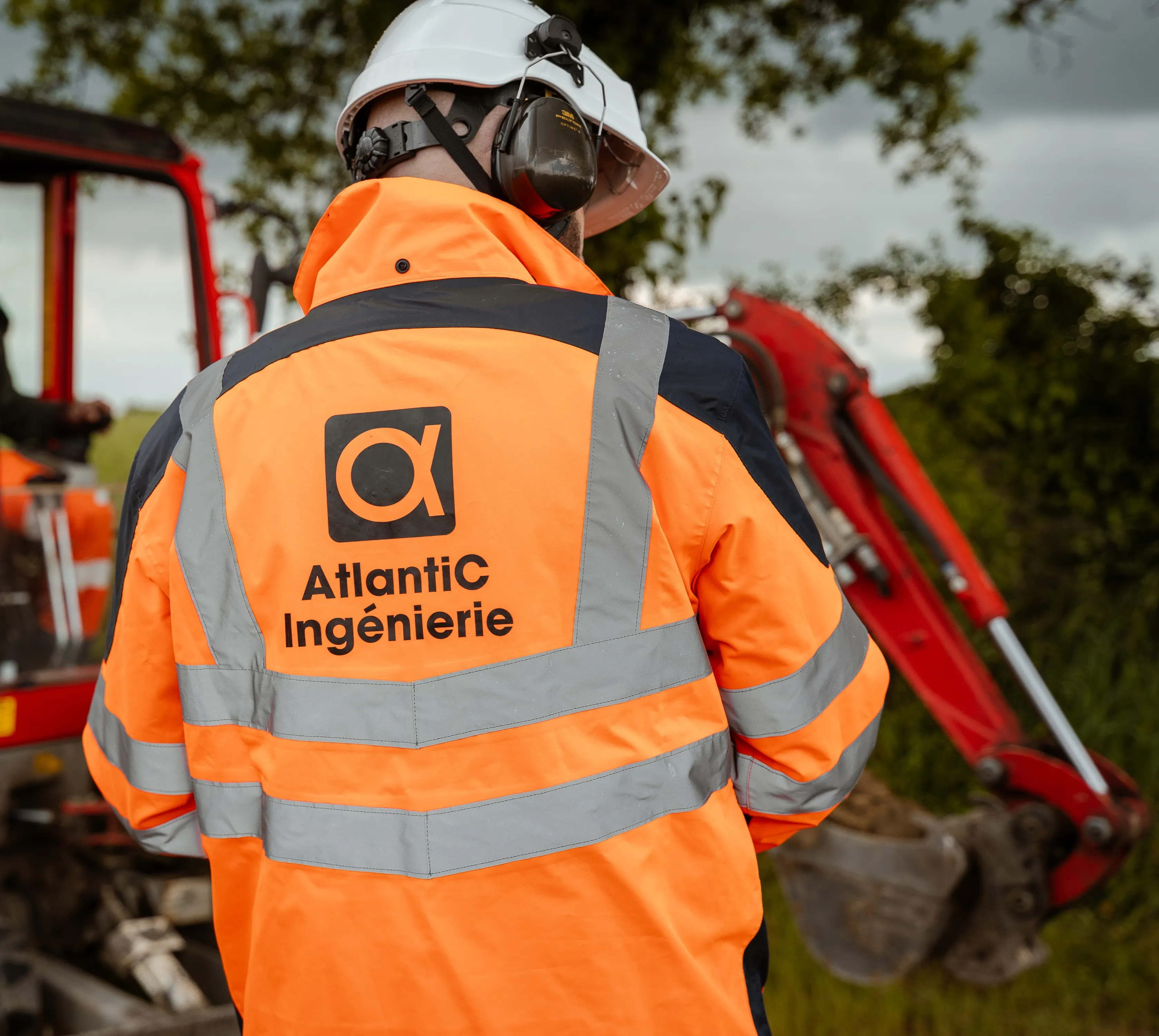 Ouvrier en veste de sécurité orange avec bandes réfléchissantes et casque blanc, portant un casque antibruit, devant une pelleteuse rouge en plein air.