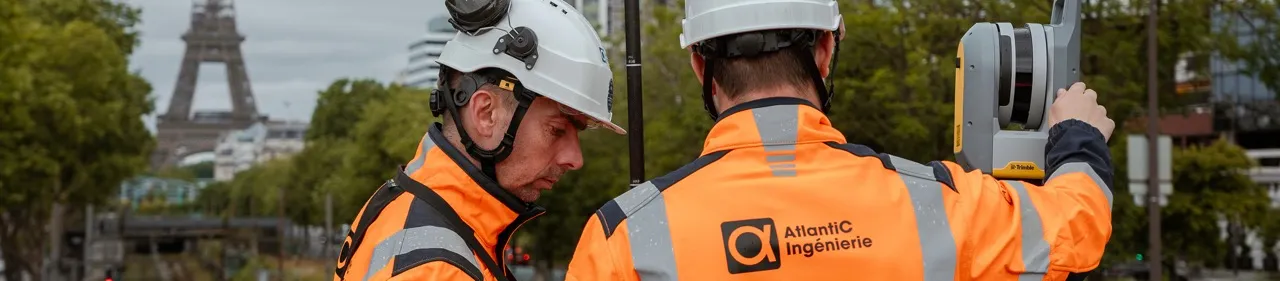 Deux ingénieurs en sécurité portant des casques et des vestes orange réfléchissantes travaillent avec un équipement de mesure Trimble en extérieur, avec la Tour Eiffel visible en arrière-plan à Paris.
