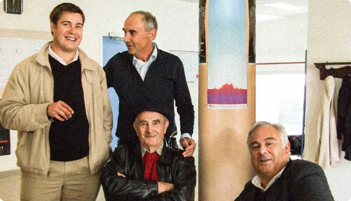 Quatre hommes souriants posant ensemble à l'intérieur devant un poteau avec une affiche colorée, certains portant des vestes et pulls.