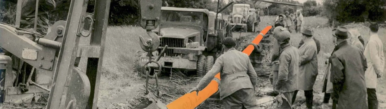 Photo en noir et blanc montrant plusieurs ouvriers et techniciens travaillant ensemble pour poser un long tuyau en terre à l'aide d'une grue et de camions.
