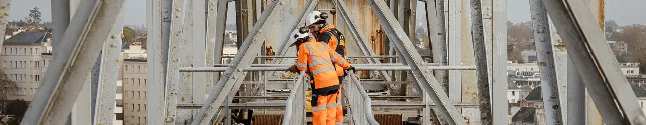 Deux travailleurs en tenue de sécurité orange et casques blancs sur une structure métallique industrielle surplombant une ville.