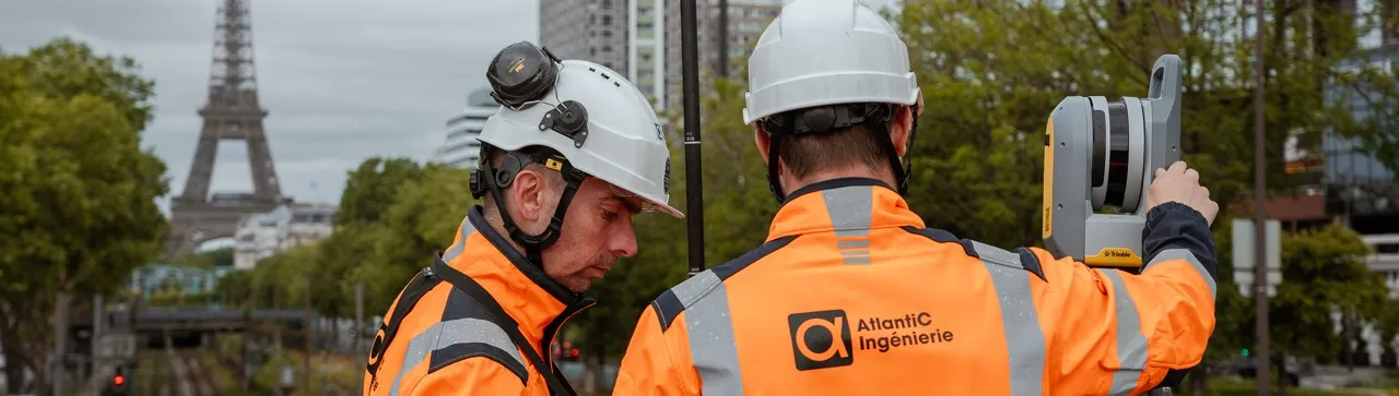 Deux ingénieurs en vestes orange réfléchissantes et casques blancs utilisent un équipement de topographie à Paris, avec la tour Eiffel visible en arrière-plan.