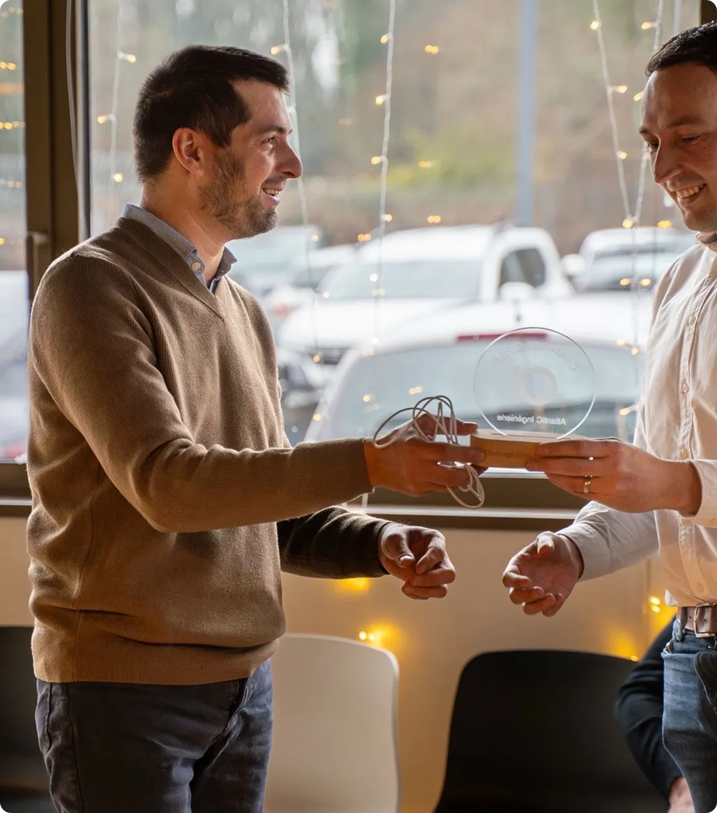 Deux hommes souriants échangent un trophée en verre et un objet rectangulaire à l'intérieur d'une pièce avec des guirlandes lumineuses et des fenêtres donnant sur un parking.