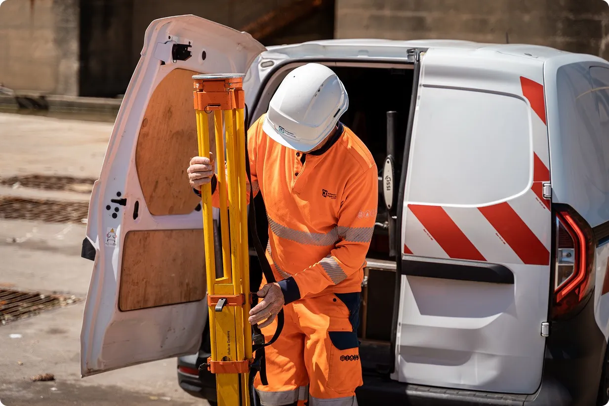 Travailleur en tenue orange de haute visibilité et casque blanc, sortant un trépied jaune d'un fourgon blanc à portes arrière ouvertes avec des bandes rouges réfléchissantes.