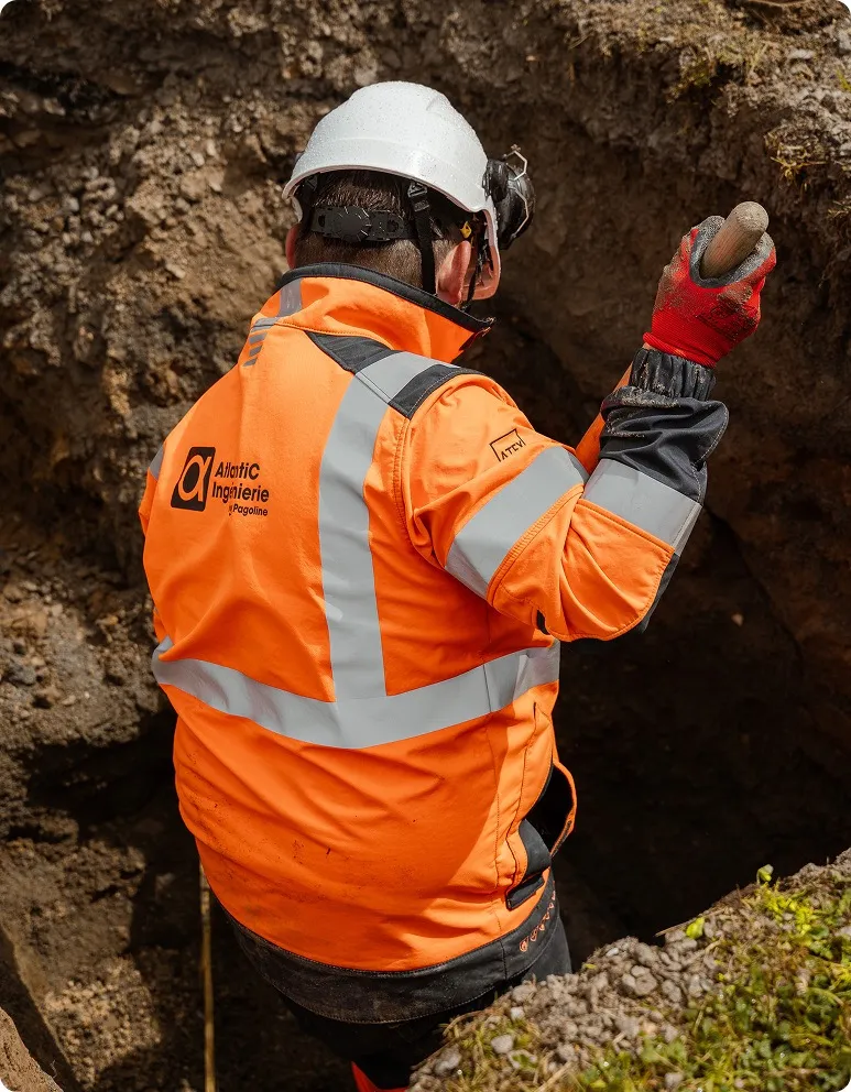 Ouvrier portant un casque blanc, une veste haute visibilité orange avec bandes réfléchissantes et des gants rouges, creusant dans la terre.