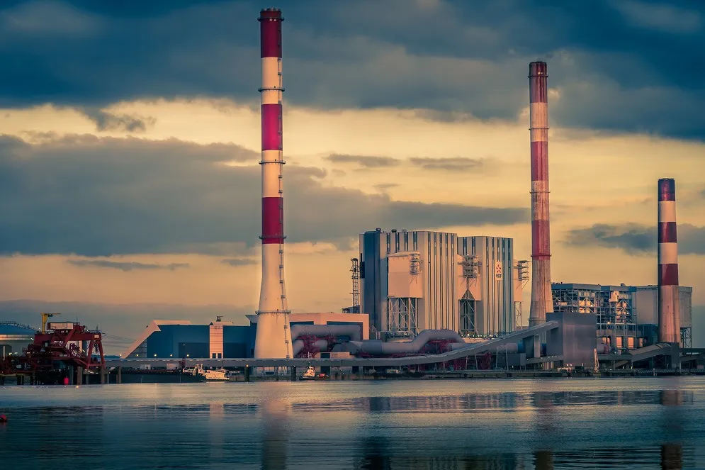 Vue d'une centrale électrique industrielle avec plusieurs cheminées rayées en rouge et blanc au bord d'une étendue d'eau sous un ciel nuageux au coucher du soleil.