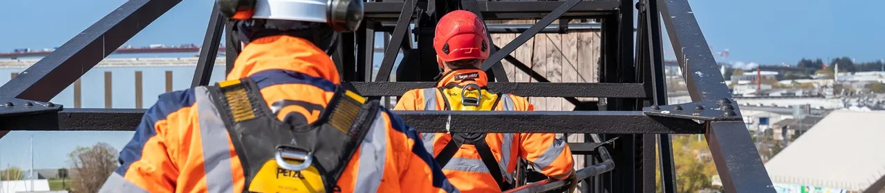 Deux ouvriers en haute visibilité orange et casques de sécurité sur une structure métallique extérieure, avec une vue urbaine en arrière-plan.
