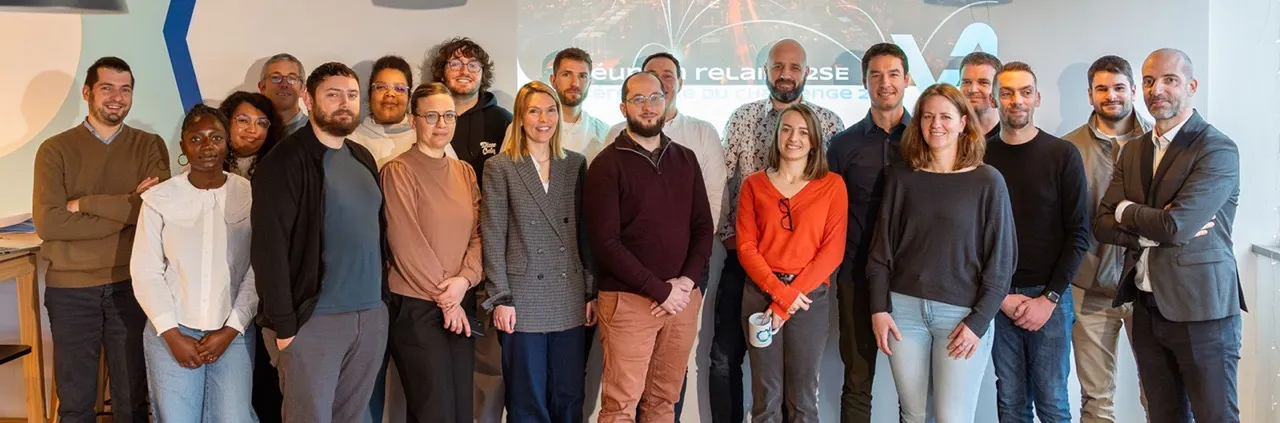 Groupe de 18 personnes debout en rangée, posant pour une photo dans un bureau lumineux avec un écran en arrière-plan affichant un logo et du texte.