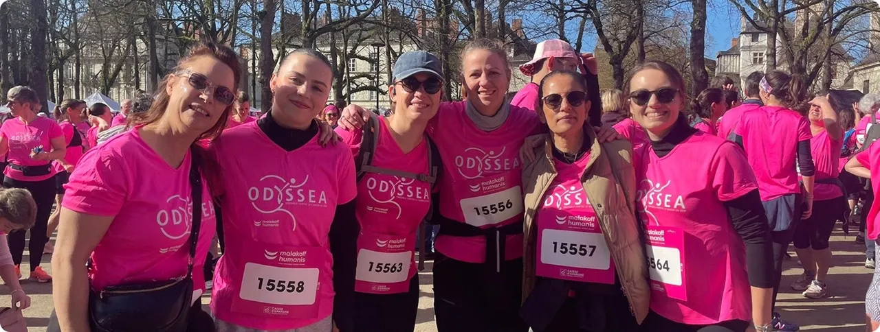 Groupe de six femmes habillées de t-shirts roses Odyssea avec des numéros de course posant ensemble dans un parc pendant une journée ensoleillée, avec d'autres participants en arrière-plan.