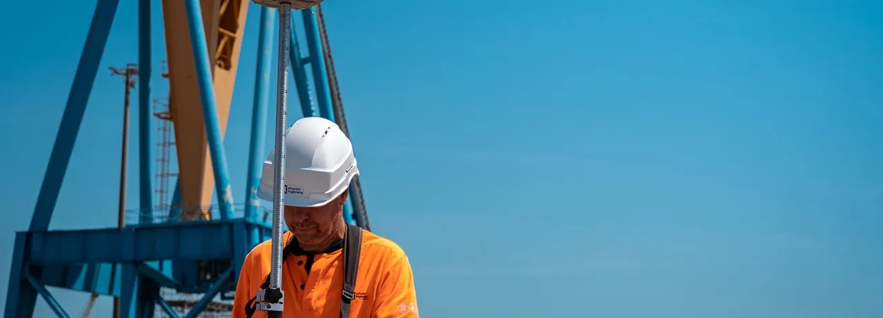 Un ouvrier portant un casque blanc et un gilet orange utilise un instrument de mesure sur un chantier avec une grande structure métallique bleue en arrière-plan sous un ciel bleu clair.