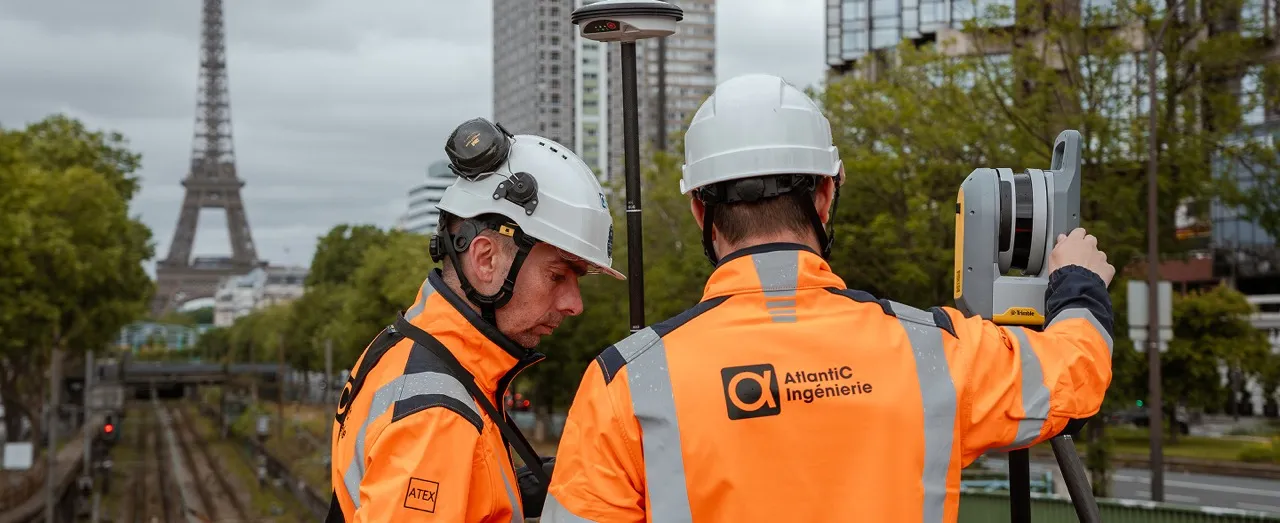 Deux ingénieurs portant des casques blancs et des vestes orange réfléchissantes opèrent un équipement de topographie près des voies ferrées, avec la tour Eiffel visible en arrière-plan à Paris.