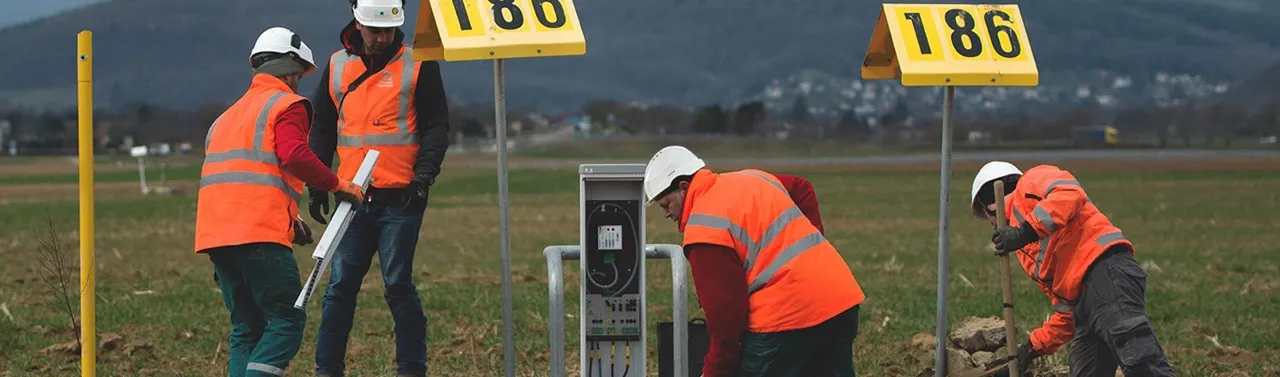 Quatre ouvriers en gilets orange et casques blancs effectuent des travaux en extérieur près d'un équipement technique. Deux panneaux jaunes avec le numéro 186 sont visibles, ainsi qu'un paysage avec des champs et des collines en arrière-plan.