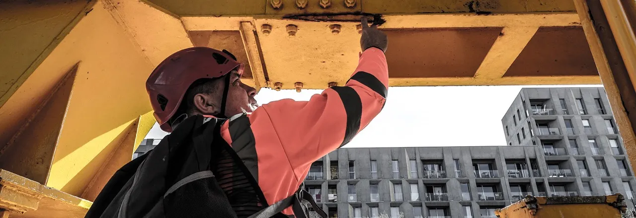 Un ouvrier portant un casque orange et une veste de sécurité inspecte une structure métallique jaune sur un chantier en milieu urbain avec des immeubles en arrière-plan.