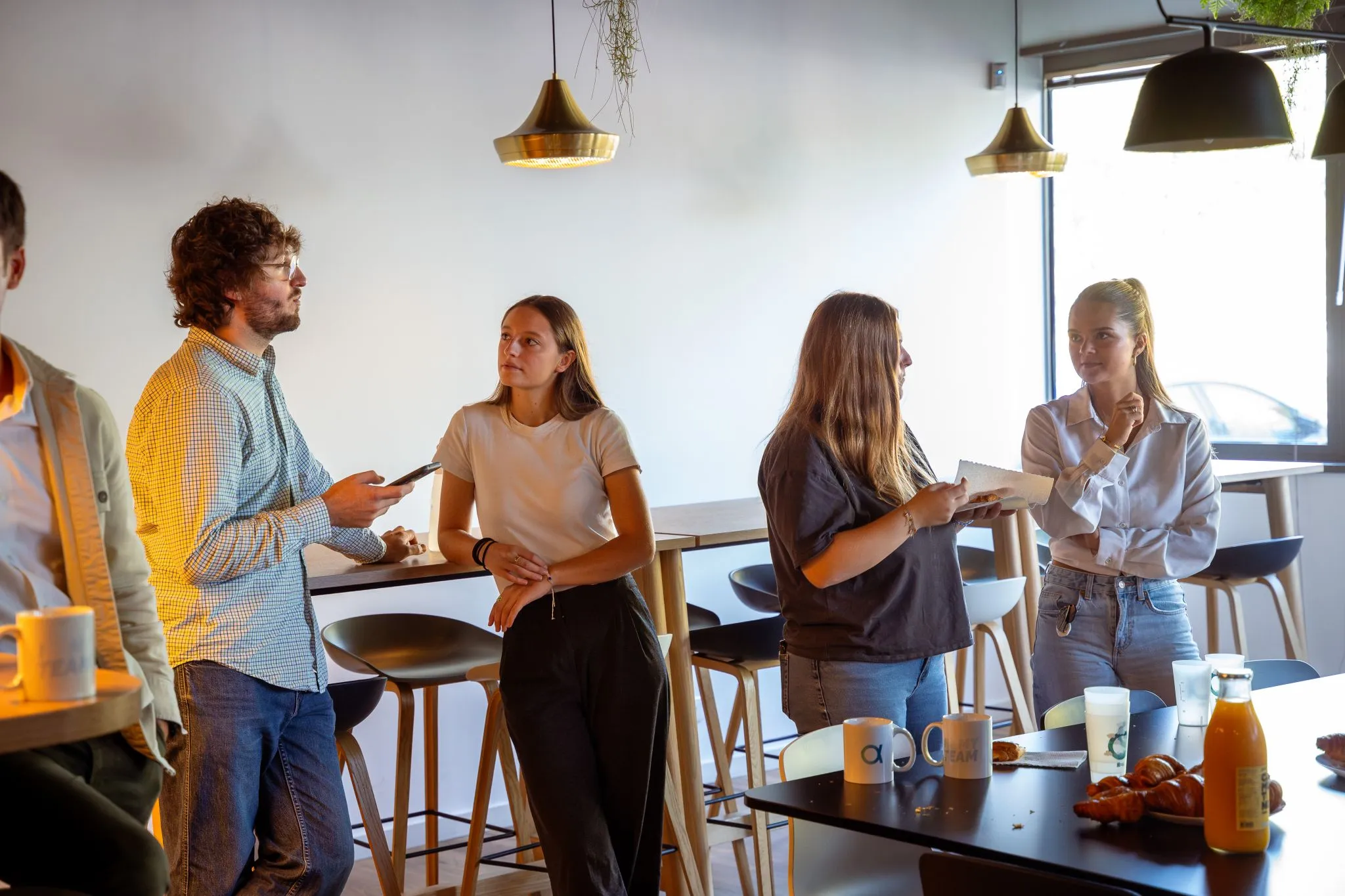 Quatre jeunes adultes discutent dans un espace de travail avec des tabourets hauts, des tasses, des croissants et une bouteille de jus sur une table.