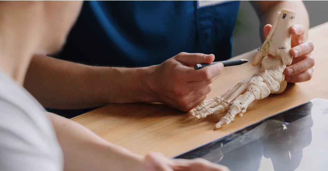 Medical professional holding a foot bone model and pointing with a pen at a patient's foot on a table.