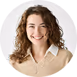 Smiling woman with curly hair wearing a beige sweater and white collared shirt against a white background.