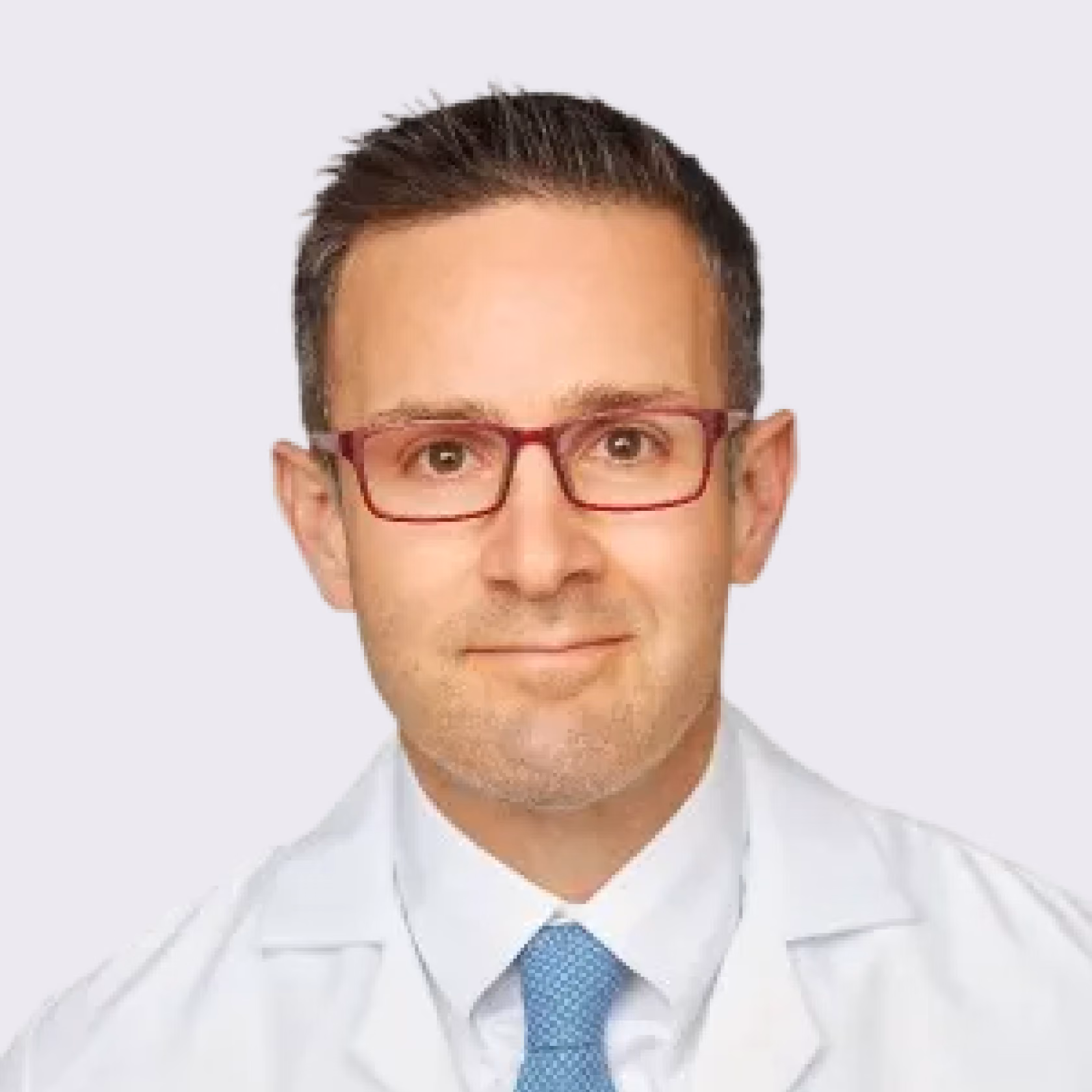 Man with short dark hair and red glasses wearing a white coat and blue tie on light background.