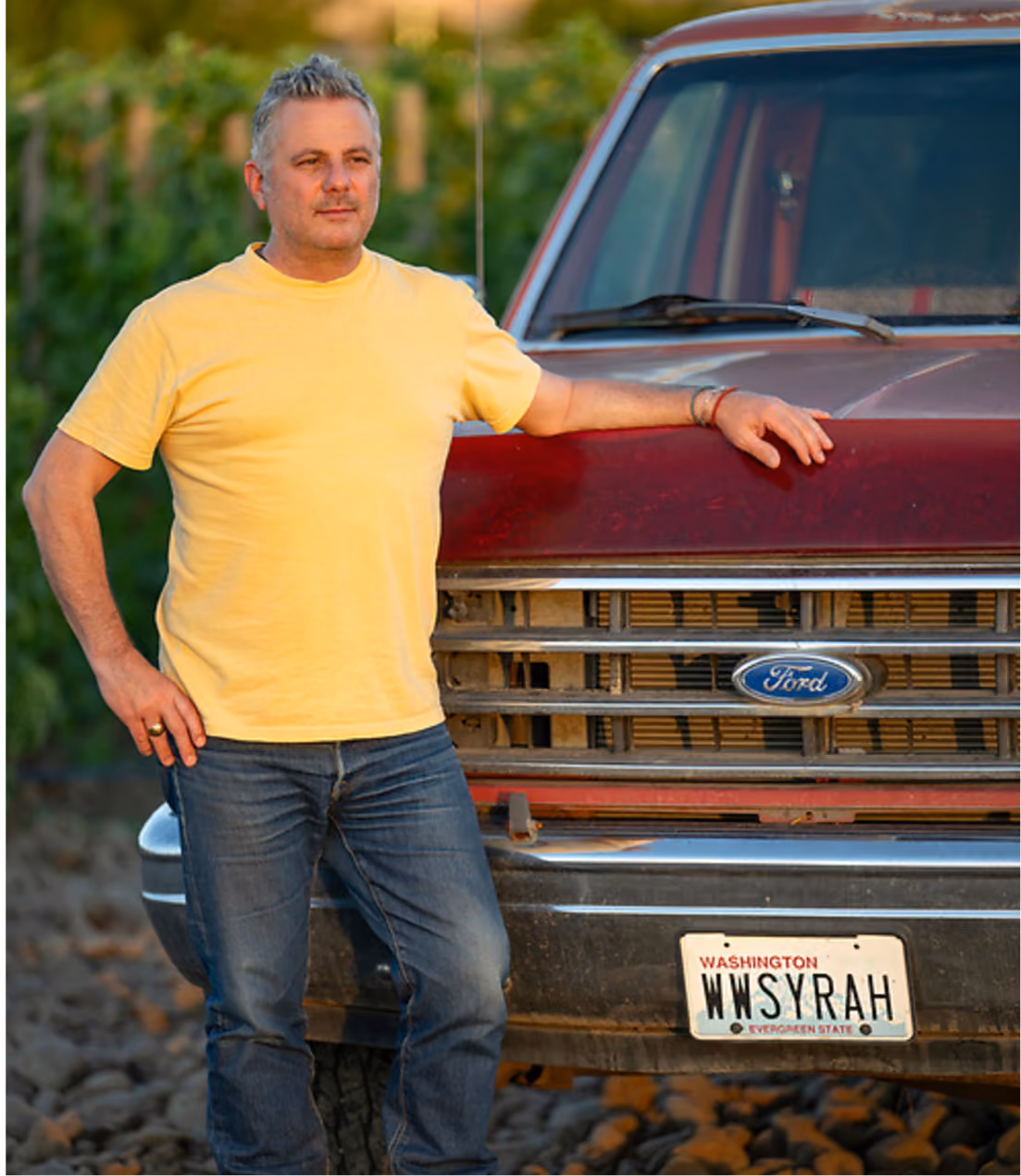 Christophe Baron in a yellow t-shirt and blue jeans leaning against the front of a red Ford truck with a Washington state license plate reading 'WWSYRAH'.