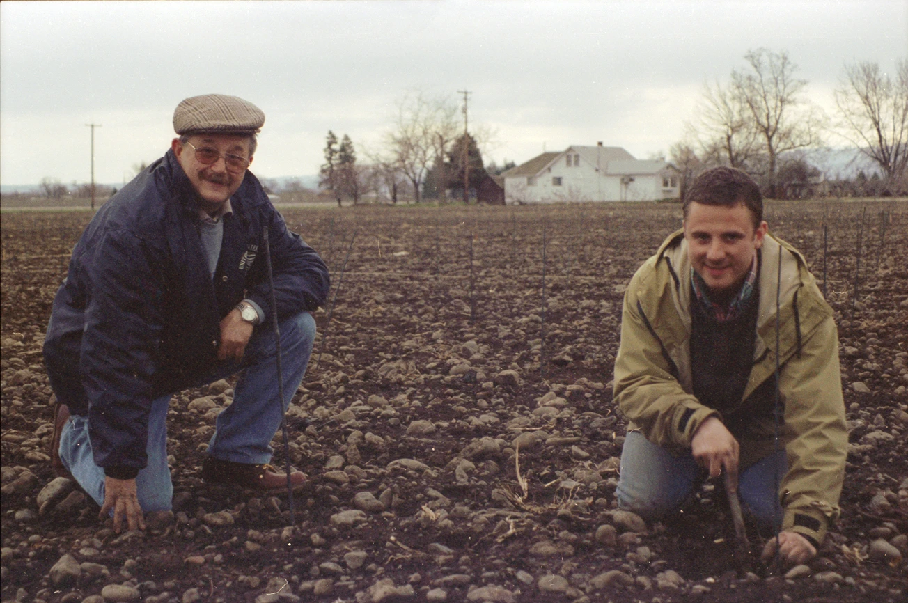 Two men kneeling in a rocky, bare field with small stakes in the soil; one man wears a jacket and hat with glasses, the other a light jacket, both smiling toward the camera, with a house and bare trees in the background under an overcast sky.