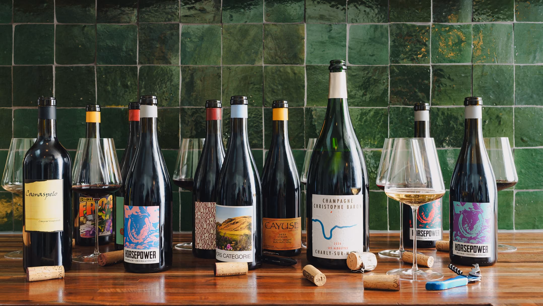 A variety of wine bottles and glasses with red and white wine arranged on a wooden surface against a green tiled wall, with corks and a corkscrew nearby.