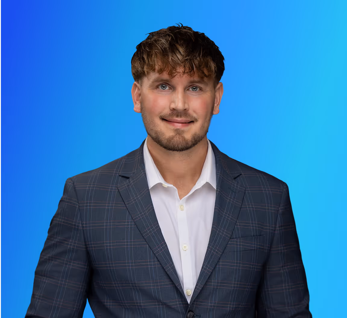 Smiling man with short brown hair and beard wearing a dark plaid suit and white shirt on blue background.
