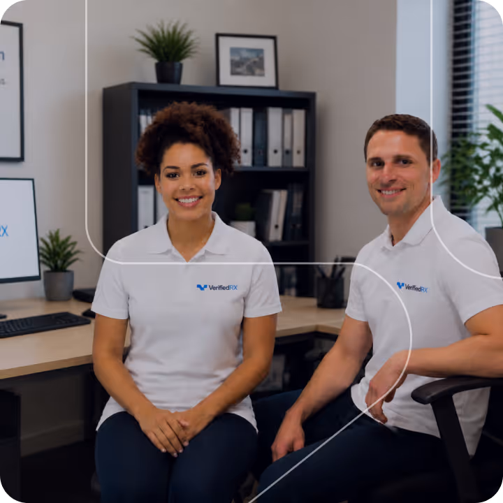 Two people wearing VerifiedRx logo shirts sitting in an office with desks, plants, and shelves behind them.
