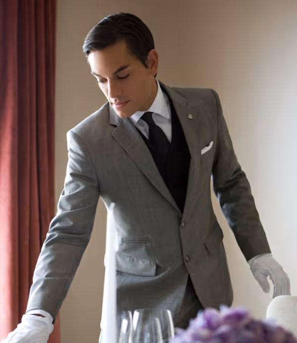 Man in gray suit and white gloves setting a table with glassware and purple flowers.