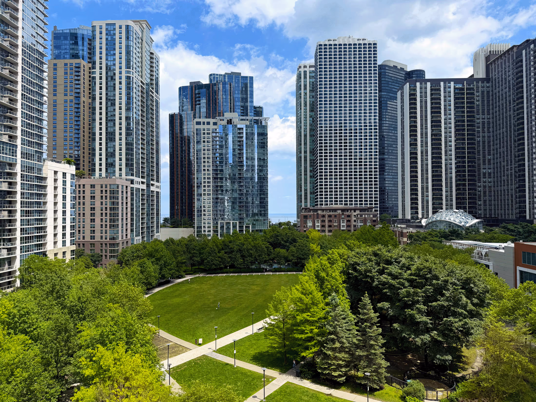 City park with green lawns and trees surrounded by tall modern skyscrapers under a partly cloudy sky.