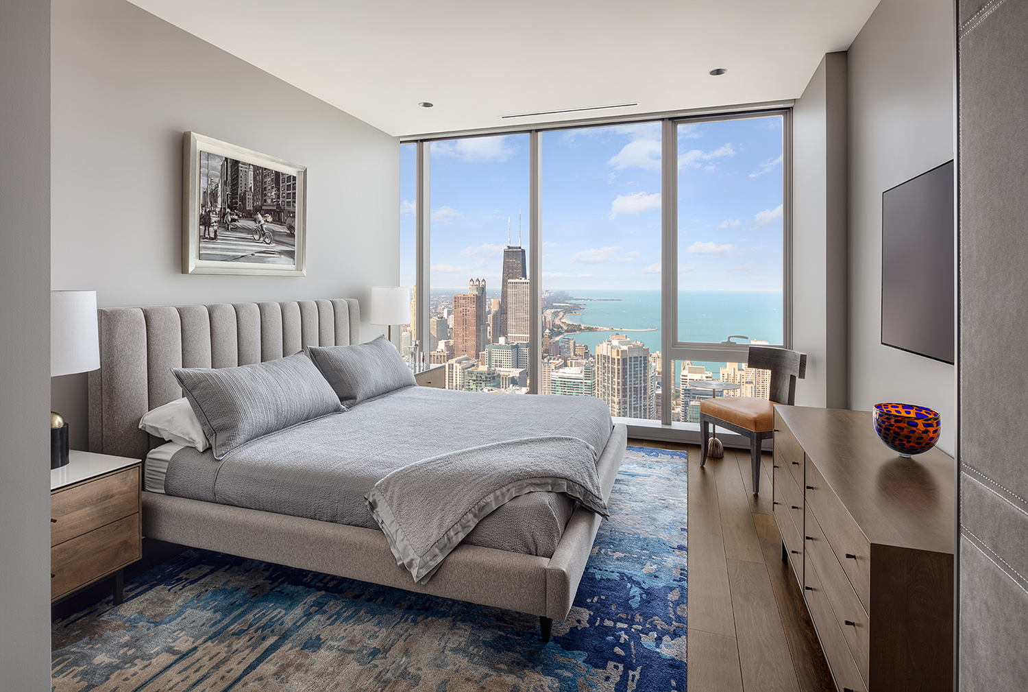 Modern bedroom with gray bed, blue rug, dresser, chair, and large windows showing city skyline and lake.