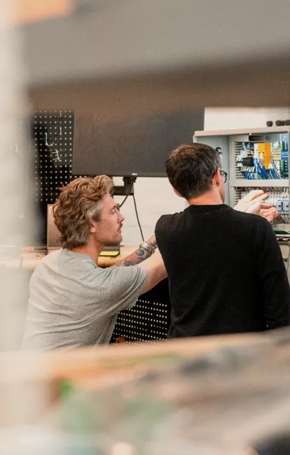 Two Podium Automation technicians examining wiring inside an open control panel at a workshop bench.