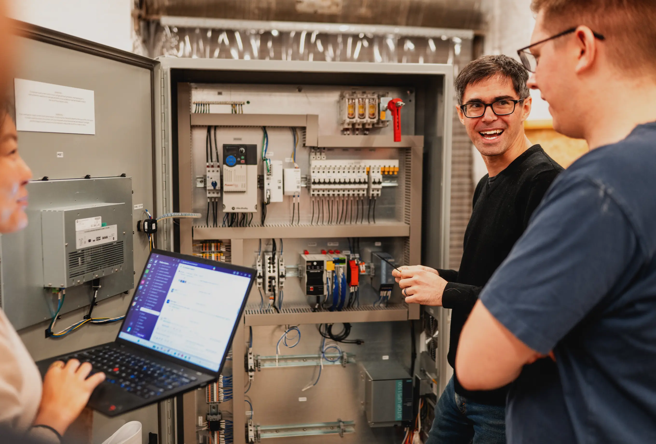 Podium Automation engineers discussing an open control panel with an Allen-Bradley PowerFlex drive, circuit breakers, and a connected laptop.