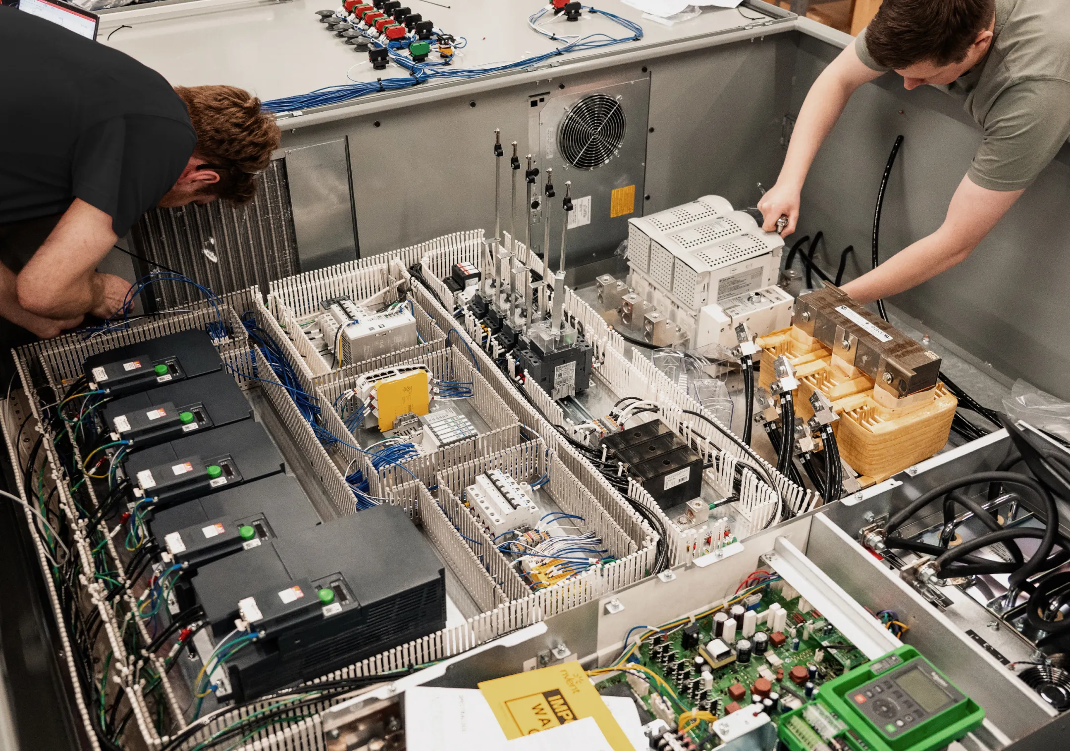 Two Podium Automation technicians wiring a large open control panel with variable frequency drives, a transformer, and wire ducts.