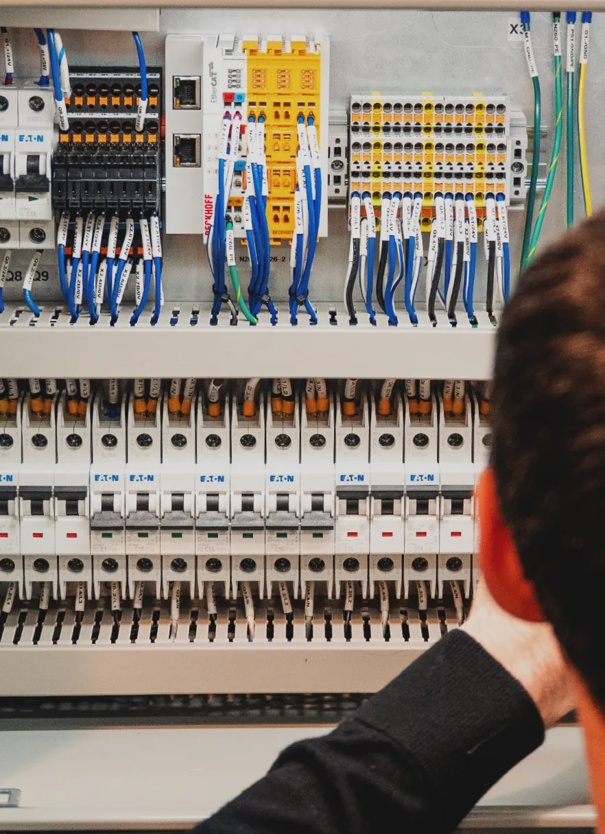 Technician inspecting a control panel with Eaton circuit breakers, Beckhoff EtherCAT I/O modules, and labeled wiring on terminal blocks.
