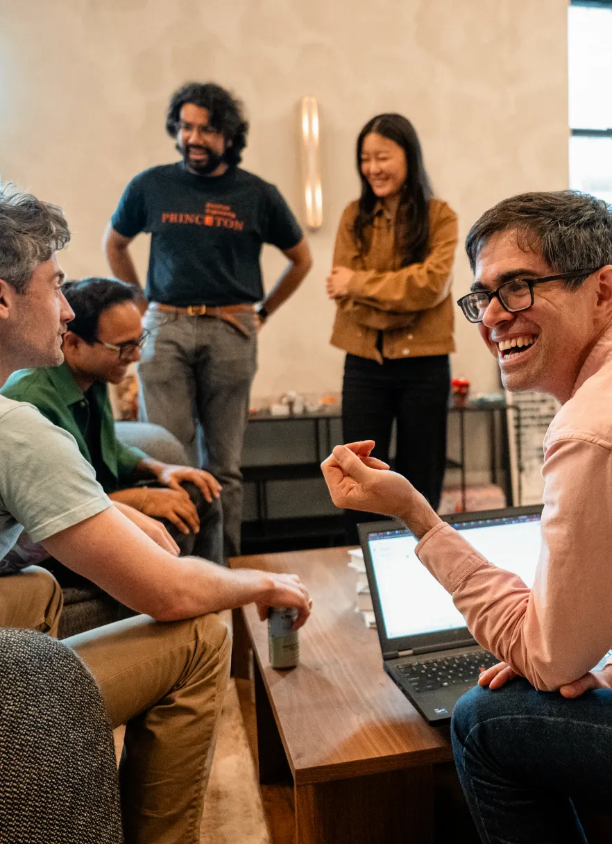 Five Podium Automation team members laughing together in a lounge area with a laptop, reflecting the team culture.