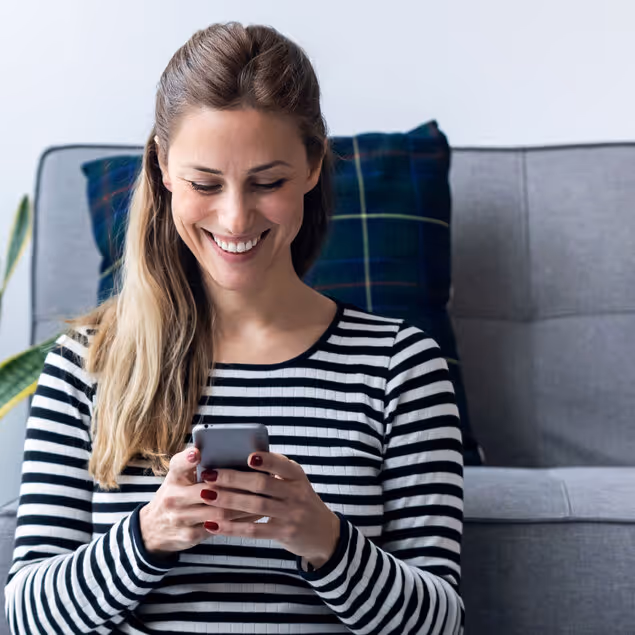 A woman with long brown hair, wearing a black and white striped shirt, smiles while looking at a smartphone in her hands.