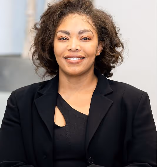 A smiling woman with shoulder-length curly hair wearing a black blazer and a black top, posing confidently with her arms crossed.